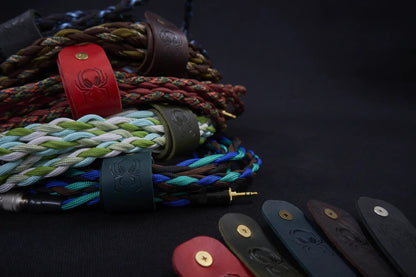 Fermata leather cable keepers wrapped around Arachne Audio braided cables with a brass 3.5mm connector, embossed spider detail.