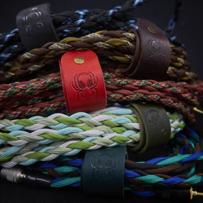 Fermata vegetable-tanned leather cable strap in red with embossed Arachne spider and brass stud, holding a coiled braided headphone cable.